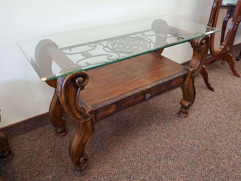 Solid wood accent table featuring beveled edge glass top, decorative bent metal support, single center drawer, scrollwork and carved accents measuring 48in W x 20-in D X 30 in H. MATCHES LOT 8.