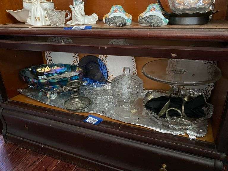 Contents of shelf: Indiana carnival glass fruit bowl, cobalt glass plate, Fostoria American covered compote, glass cakestand, spoon napkin rings, misc