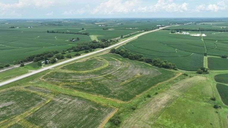 Elevated drone photo of a 27-acre Washington County, Iowa parcel for sale, showing concentric contour-planted fields, grass waterways, and long frontage on a paved highway.