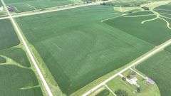 Aerial drone photograph of a 106-acre Washington County, Iowa row-crop farm for sale—lush cornfields shaped by gentle contour farming and bordered on two sides by paved roads.