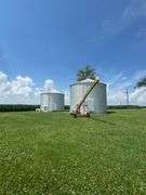 Drone view of two silver, corrugated-steel grain bins on a neatly mown farmyard, framed by tall cornfields—part of a 42-acre Washington County, Iowa farm for sale.