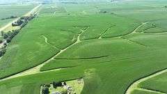 Aerial view of a 70-acre Washington County, Iowa farm for sale—lush cornfields shaped by branching grass waterways, bordered by a paved highway and timbered rail corridor.
