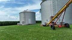 Drone view of two silver, corrugated-steel grain bins on a neatly mown farmyard, framed by tall cornfields—part of a 42-acre Washington County, Iowa farm for sale.