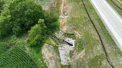 Aerial close-up of a new concrete culvert with riprap stabilizing a small creek as it passes under a paved highway, bordered by crop rows and trees—part of a 27-acre tract for sale in Washington County, Iowa.