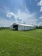 Drone view of a weathered open-front machine shed and two silver grain bins set amid lush cornfields—part of a 42-acre Washington County, Iowa farm for sale.