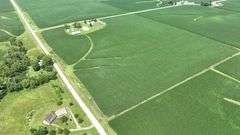 Aerial drone photograph of a 42-acre Washington County farmland tract for sale, showing lush row-crop fields plus two white machine sheds and two round grain bins along a paved road.