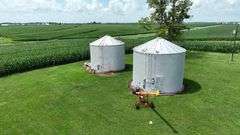 Drone view of two silver, corrugated-steel grain bins on a neatly mown farmyard, framed by tall cornfields—part of a 42-acre Washington County, Iowa farm for sale.