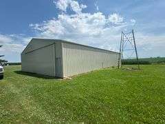 Ground-level drone view of a tan steel machine shed on a well-kept farmyard, with grain bins, cornfields, and open sky—part of a 42-acre Washington County property for sale.