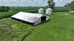 Drone view of a weathered open-front machine shed and two silver grain bins set amid lush cornfields—part of a 42-acre Washington County, Iowa farm for sale.