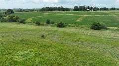 Mid-elevation drone view of a 30-acre Washington County tract for sale, featuring rolling prairie grass in the foreground, a brush-lined drainage swale, and row-crop fields with a distant farmstead.