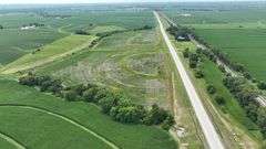 Elevated drone photo of a 27-acre Washington County, Iowa parcel for sale, showing concentric contour-planted fields, grass waterways, and long frontage on a paved highway.