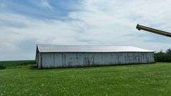 Drone view of a weathered open-front machine shed and two silver grain bins set amid lush cornfields—part of a 42-acre Washington County, Iowa farm for sale.