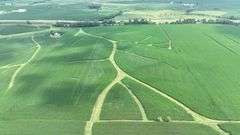 Aerial drone view of 70 contiguous acres of Washington County cropland for sale—lush soybean fields patterned with branching grass waterways and bordered by a tree-lined rail spur.