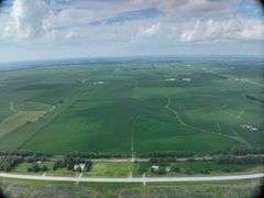 Aerial view of a 72-acre Washington County, Iowa row-crop field for sale—uniform green soybeans on gently rolling ground bordered by grass waterways and a nearby paved highway.