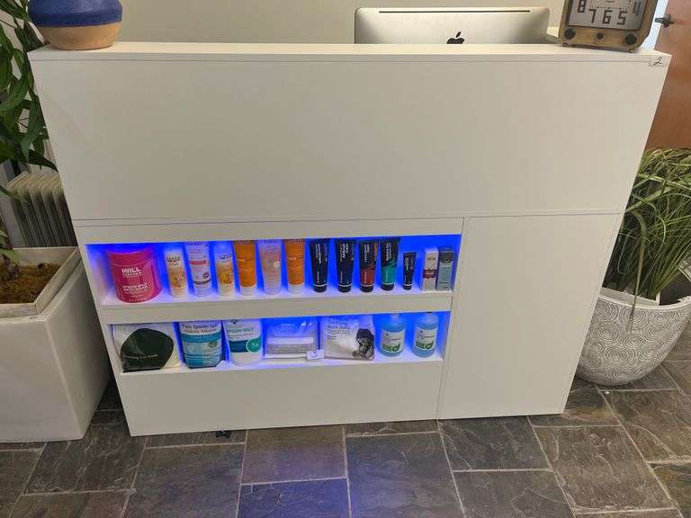 modern white reception counter with illuminated shelves displaying various hair care products. The sleek design and organized product layout create a clean, professional look suitable for a beauty salon or wellness clinic.