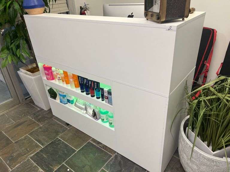 modern white reception counter with illuminated shelves displaying various hair care products. The sleek design and organized product layout create a clean, professional look suitable for a beauty salon or wellness clinic.