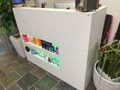 modern white reception counter with illuminated shelves displaying various hair care products. The sleek design and organized product layout create a clean, professional look suitable for a beauty salon or wellness clinic.